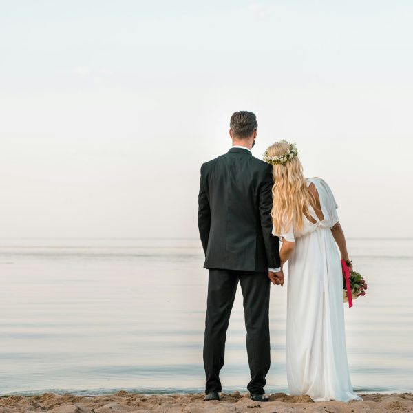 back view of wedding couple standing on beach with wedding bouquet and looking at sea