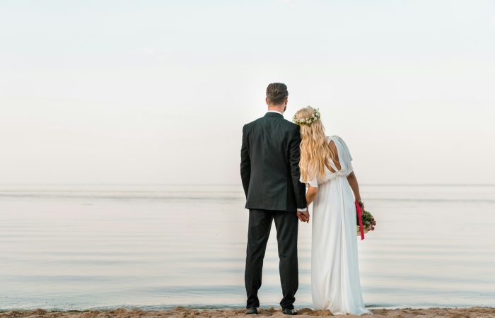 back view of wedding couple standing on beach with wedding bouquet and looking at sea